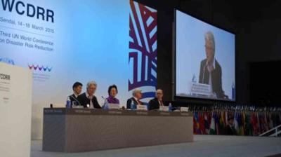 A panel of five people sits at a long table at the Third UN World Conference on Disaster Risk Reduction, with a large screen displaying a speaker behind them.