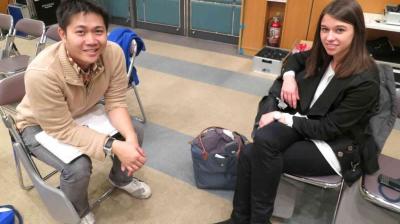 Two people are seated on chairs in a room, facing the camera. One has papers on their lap; both are casually dressed. A fire extinguisher is visible in the background.