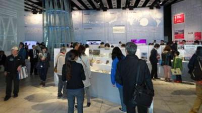 People explore exhibits in a well-lit museum or gallery space with informational displays on walls and in glass cases.