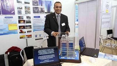 A man stands behind a table displaying a small architectural model, accompanied by two open laptops and informational posters in the background at a booth.
