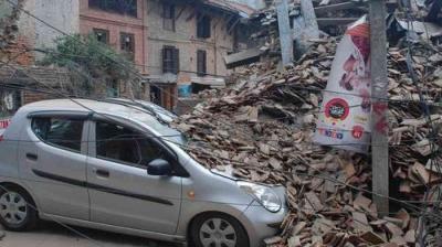 A silver car is parked beside a large pile of rubble from a collapsed building, with fallen debris and power lines surrounding the area.