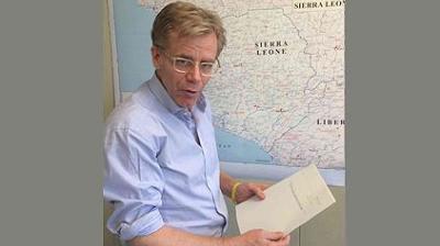 A person in a light blue shirt stands holding a document in front of a map showing parts of West Africa, including Sierra Leone and Liberia.
