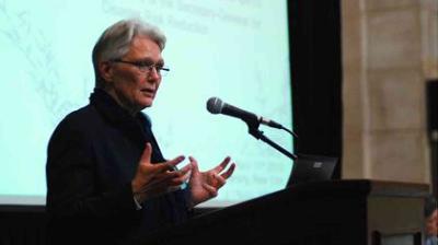Person speaking at a podium with a microphone, gesturing with hands. A projected presentation screen is visible in the background.