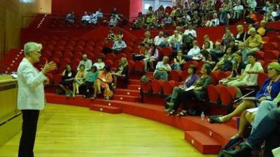 A person stands and speaks to an audience seated in red chairs in a tiered lecture hall.