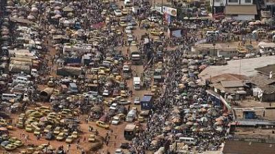 A busy urban street is filled with numerous vehicles, people walking, and market stalls, creating a crowded scene with heavy traffic and bustling activity.