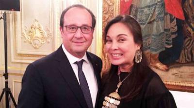 A man in a suit stands next to a smiling woman in a black outfit with large earrings, inside an ornately decorated room.