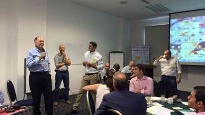 Several people are gathered in a conference room; one man is speaking into a microphone while others listen, with a projector screen and whiteboard in the background.