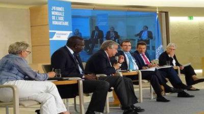 A panel of six people seated on stage during a discussion. A sign in the background reads "Journée Mondiale de l’Aide Humanitaire.