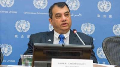 A man speaking at a conference with a UN-themed background, sitting at a table with a nameplate and microphone.