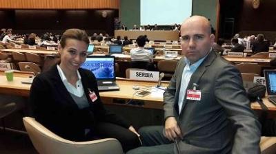 Two individuals seated in a conference room with laptops, surrounded by people and nameplates, including one labeled "Serbia.
