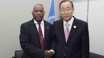 Two men in suits shake hands in front of a United Nations flag in an office setting.