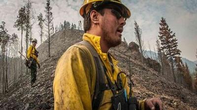 Two firefighters in yellow protective gear stand on a rocky, forested hillside, surveying the area. One is in the foreground, and the other is further back with trees and mountains in view.