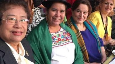 Four women sitting together, smiling at the camera. One woman wears traditional clothing with a green shawl and black hat, while the others are dressed in modern attire.