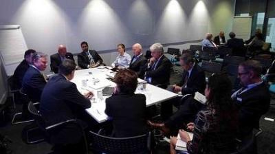 Group of people in formal attire seated around a conference table in a meeting room with a whiteboard and a presentation board in the background.