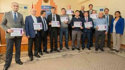 A group of fourteen people stand indoors, several holding certificates, posing for a photo during an award or recognition event.