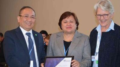 Three adults in business attire stand together indoors, with one person holding a framed certificate and all smiling at the camera.