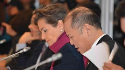 Two people are sitting side by side, one reading from a paper, in a conference setting with microphones in front of them.