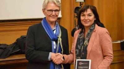 Two women shaking hands while holding a plaque in a wood-paneled room. One wears a blue scarf, and the other, a pink blazer.