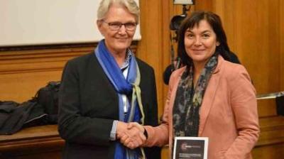 Two women are shaking hands; one is holding a plaque. They are standing in a wood-paneled room with a projector screen in the background.