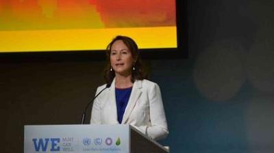 A woman in a white blazer stands at a podium, speaking at an event with a colorful background screen behind her.