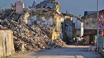 A damaged building with collapsed walls and debris scattered on the street after an apparent earthquake; two people walk in the distance.