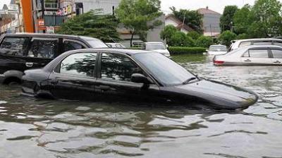 Cars partially submerged in a flooded street, with trees and buildings in the background.