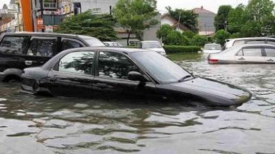 Several cars are partially submerged in floodwater on a city street, with buildings and trees visible in the background.