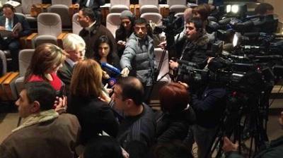 A group of reporters and camera operators surround a person speaking at a press event in an auditorium with empty seats in the background.