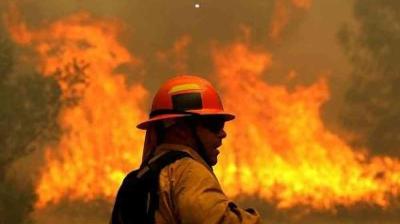 A firefighter in safety gear stands in front of a large wildfire, with flames and smoke visible in the background.