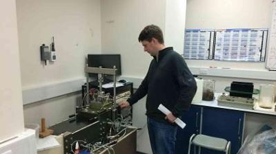 Person in a lab, operating scientific equipment on a desk, with charts and shelving in the background.