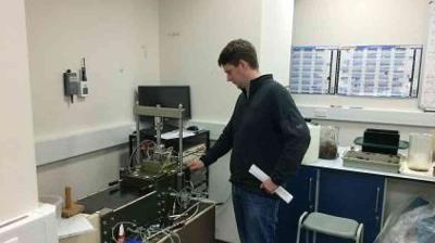 A person stands next to laboratory equipment, adjusting a machine with wires and tubes, in a room with a computer and charts on the wall.
