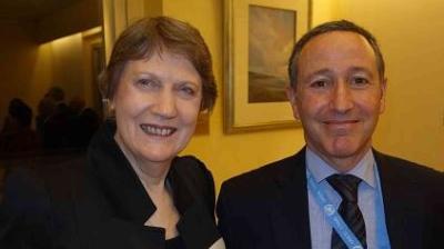 Two people, a woman and a man, smiling in a warmly lit room with framed artwork on the walls. The man is wearing a lanyard.