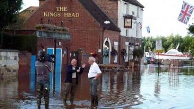 A news reporter interviews a man in front of a flooded pub, with the water reaching their knees. A camera operator films the scene. Floodwater surrounds the building.