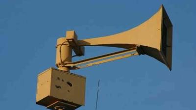 A large, beige outdoor loudspeaker mounted on a pole against a clear blue sky.