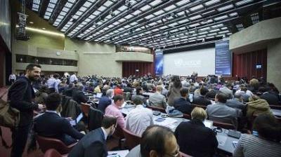 A large conference room filled with seated attendees facing a stage with a screen displaying a logo.