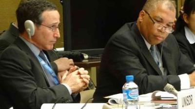 Two men in suits seated at a conference table, one wearing headphones and holding his hands together, the other writing notes with a bottle of water nearby.