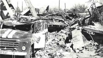 A damaged truck is parked amid rubble and collapsed concrete structures in a heavily destroyed area.