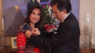 A man in a suit pins an award or decoration onto a woman wearing a patterned dress; both are standing indoors with a painting and flowers in the background.