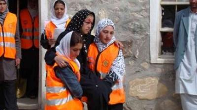 Several women wearing orange safety vests assist and support another woman outside a stone building.