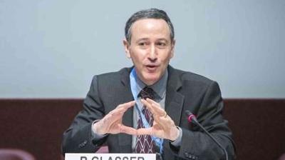 A man in a suit speaks into a microphone, using hand gestures for emphasis.