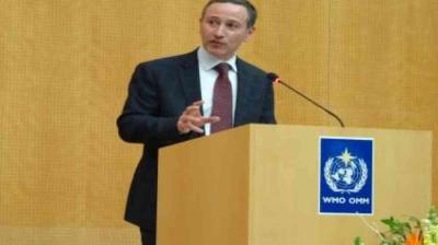 A man in a suit speaks at a podium with a microphone; the podium displays the WHO emblem against a wood-paneled background.