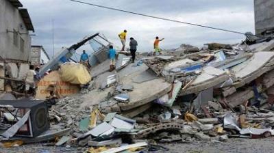 Several people stand and search on top of the rubble of collapsed buildings after an earthquake, with debris scattered around.