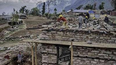 Construction workers wearing helmets build a stone house in a rural, mountainous area, with scattered debris and unfinished structures around them.