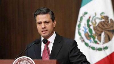 A man in a suit speaks at a podium with microphones, in front of a partial view of the Mexican flag.