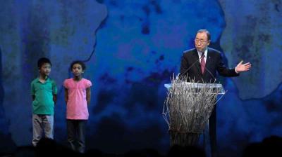 A man speaks at a podium decorated with branches, while a boy and a girl stand on stage beside him against a blue background.