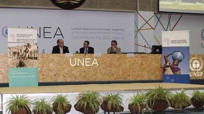 Three people sit at a panel table at the UNEA event. Banners on the stage feature environmental themes. Potted plants are arranged in front of the table.