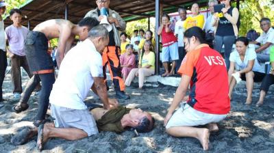 A group of people assists a man lying on the ground at a beach, while bystanders watch in the background.