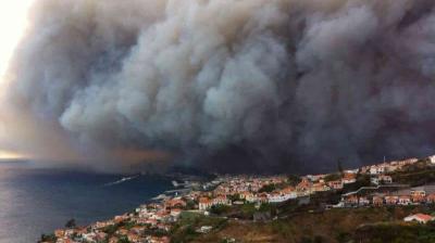 Thick smoke from a large wildfire engulfs the sky above a coastal town with red-roofed buildings near the sea.