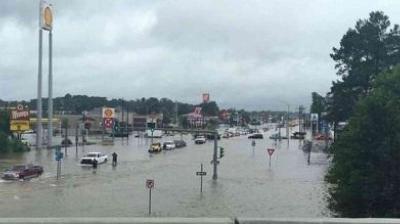 A flooded street with partially submerged cars and road signs; businesses and trees are visible in the background under a cloudy sky.