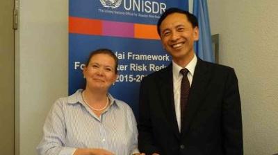 Two people stand in front of a UNISDR banner, smiling and shaking hands at an event related to disaster risk reduction.
